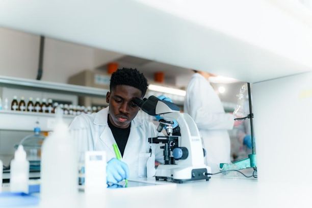 Young male scientist wearing lab coat and gloves using microscope and taking notes during scientific experiment in modern laboratory with colleague out of focus in background; Shutterstock ID 2588932423; purchase_order: Bynder library; job: ; client: ; other: 
